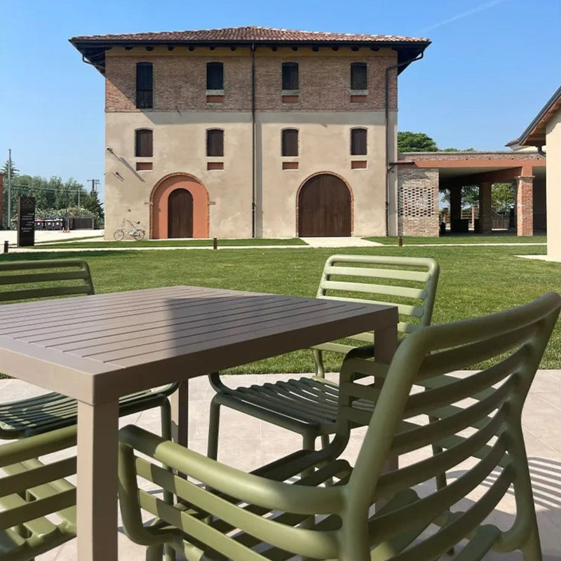 Modern outdoor dining set featuring a brown resin table and green resin armchairs, placed on a tiled patio overlooking a lush lawn and a rustic villa, evoking a serene countryside ambiance.