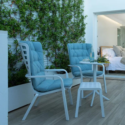 Pair of white resin lounge chairs with blue cushions, accompanied by a matching side table and stool, set on a wooden patio surrounded by vibrant green vines, embodying the essence of outdoor living.