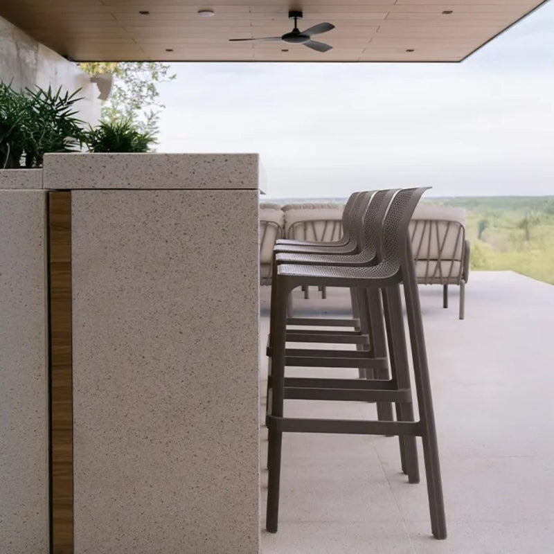 Sleek resin bar stools in Tortora positioned at a textured stone countertop, set in a modern outdoor space with lush greenery and a serene open view, embodying the essence of outdoor living.