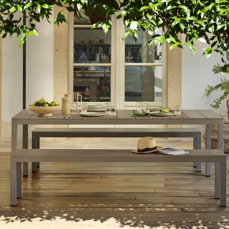 A brown Nardi Rio outdoor bench with a matching table set with fruit and cutlery, placed on a wooden deck surrounded by lush greenery. In the background, a kitchen window adds to the seamless flow between indoor and alfresco living, creating a relaxed and inviting outdoor space.