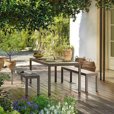 A brown Nardi Rio outdoor bench with a matching table set with fruit and cutlery, placed on a wooden deck surrounded by lush greenery. In the background, a kitchen window adds to the seamless flow between indoor and alfresco living, creating a relaxed and inviting outdoor space.