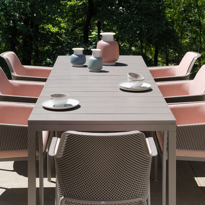 Alfresco furniture set up featuring a grey Nardi Rio extension table, complemented by seven grey and pink-accented chairs, set with cutlery in a cozy backyard setting.