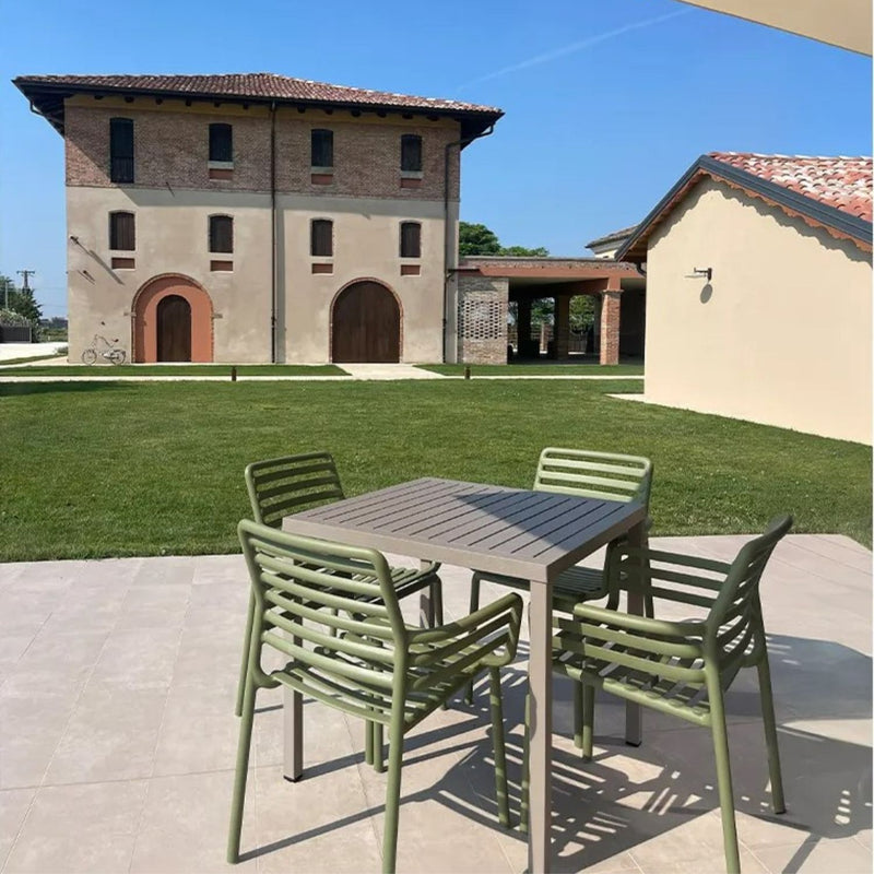 Modern outdoor dining set featuring a brown resin table and green resin armchairs, placed on a tiled patio overlooking a lush lawn and a rustic villa, evoking a serene countryside ambiance.