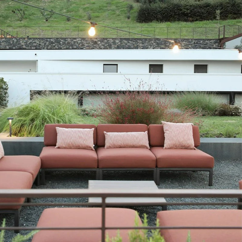 Three Nardi Maximo Outdoor Armless Chairs in Terra Russet with matching pillows, arranged side by side in a modern outdoor space. The setting features greenery, string lights, and a sleek white building in the background.
