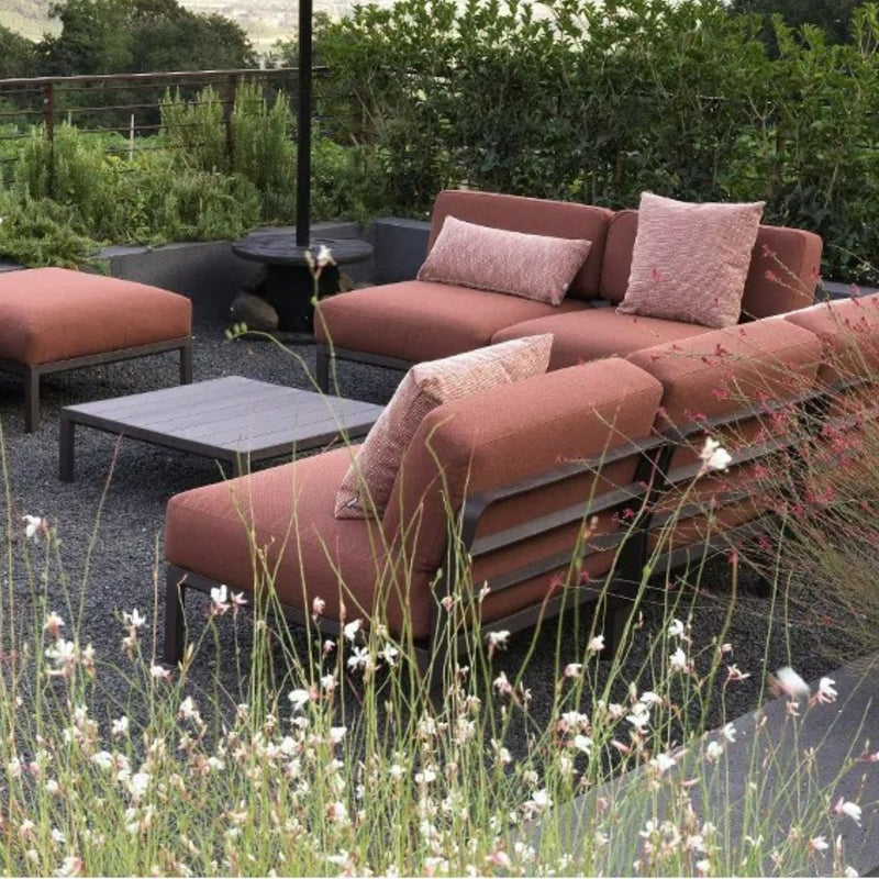 Three Nardi Maximo Outdoor Armless Chairs in Terra Russet with matching pillows, arranged around a low brown coffee table, framed by blooming white and pink flowers, lush green shrubs, and a scenic hillside backdrop, creating a serene and inviting atmosphere.