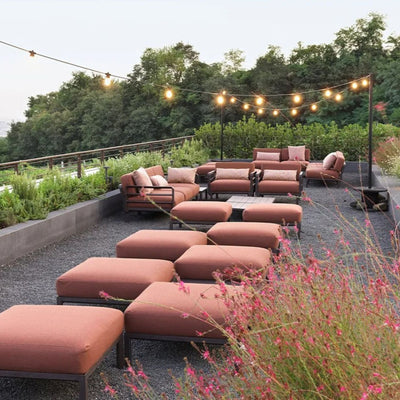 Outdoor seating arrangement featuring terra russet ottomans and armchairs with matching cushions, surrounded by lush greenery and delicate pink flowers, under warm string lights, capturing the charm of evening outdoor gatherings.