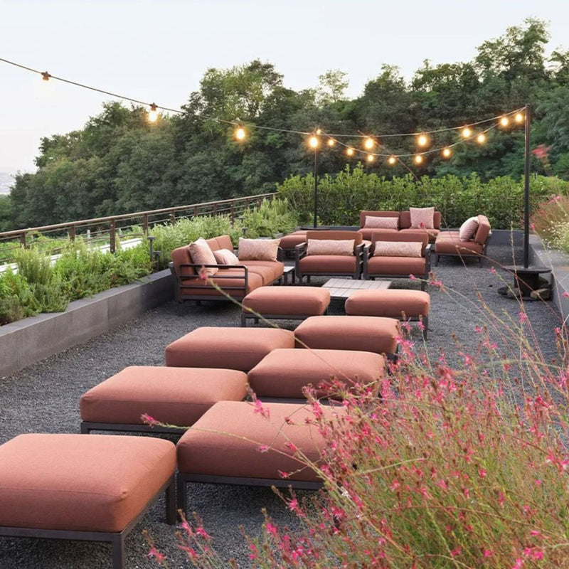 Outdoor seating arrangement featuring terra russet ottomans and armchairs with matching cushions, surrounded by lush greenery and delicate pink flowers, under warm string lights, capturing the charm of evening outdoor gatherings.