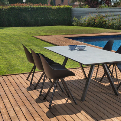 6 black resin 'Neverland' outdoor dining chairs with curved backrests, aluminium frames, and dark grey seat cushions surround a matching ceramic and aluminium outdoor dining table in someone's backyard area by a pool, with greenery in the background.