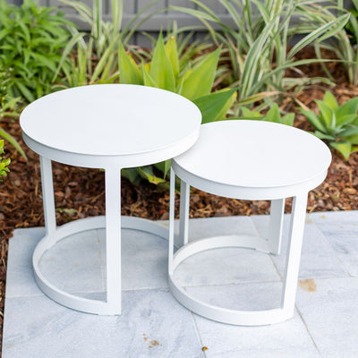 Two white round aluminium 'Neverland' outdoor side tables nestled together in a sunny outdoor area on stone tiles. The background features lush greenery, suggesting a garden setting.