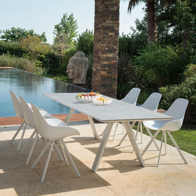 A modern outdoor dining area featuring a sleek, rectangular white ceramic dining table with splayed aluminium legs. The table is set by a pool with matching white chairs. The background features a sculpture, and the table is adorned with fruit.