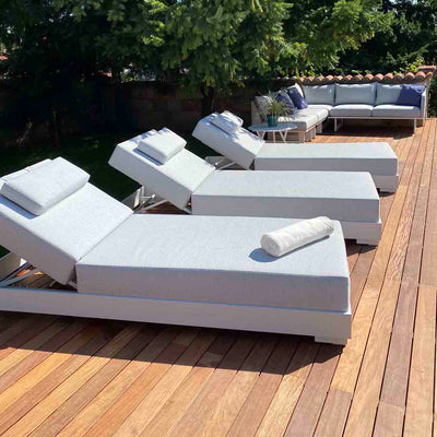Three white & light-grey aluminium 'Skyline' outdoor sun loungers with headrests slightly elevated, set on an outdoor wooden deck. One lounger has a rolled-up towel on it. In the background, there's an outdoor lounge with a coffee table and greenery surrounding the area.
