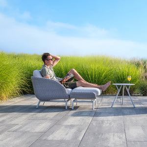 A man relaxing outdoors in a Sorrento armchair and ottoman by Couture Jardin, surrounded by lush green grass. The armchair features a sleek woven rope design with plush white cushions, paired with a small side table holding a glass of orange juice.