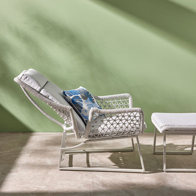 A light-grey modern outdoor furniture balcony setup made from rope features an adjustable sleek chair, reclined backrest, and a matching footrest. The furniture is viewed from the side in a room with a green wall with the sun shining in. On the chair is a decorative blue floral throw pillow.