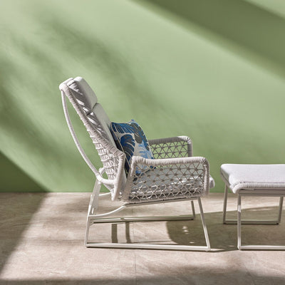 A light-grey modern outdoor furniture balcony setup made from rope featuring an adjustable sleek chair, and a matching footrest. The furniture is viewed from the side in a room with a green wall with the sun shining in. On the chair is a decorative blue floral throw pillow.