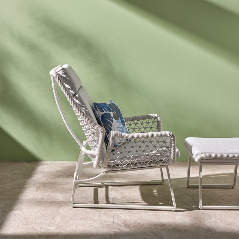 A light-grey modern outdoor furniture balcony setup made from rope featuring an adjustable sleek chair, and a matching footrest. The furniture is viewed from the side in a room with a green wall with the sun shining in. On the chair is a decorative blue floral throw pillow.