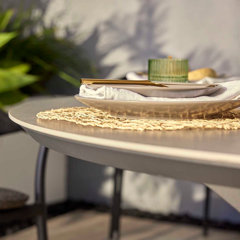 Close-up front view of the Trivento alfresco furniture round table with fruit and drinks, Neolith® ceramic surface, and blurred greenery with a grey wall in the background.