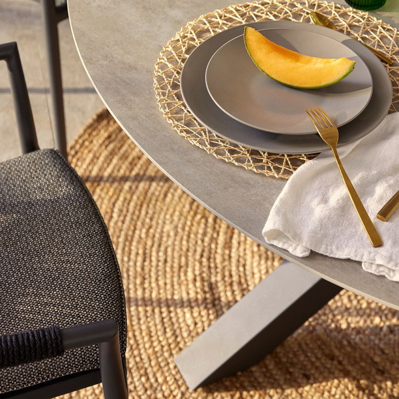 A close-up, top view of a modern outdoor dining setting featuring a round charcoal ceramic table with a minimalist design. A rope chair sits beside the table, and on top of the table is a plate served with rockmelon and a golden fork.