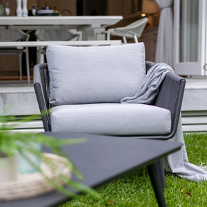 A front view of a stylish outdoor armchair with a charcoal finish, featuring a woven rope design on the backrest and seat. The chair has a sturdy, minimalist frame and is placed in an outdoor setting with greenery in the background.