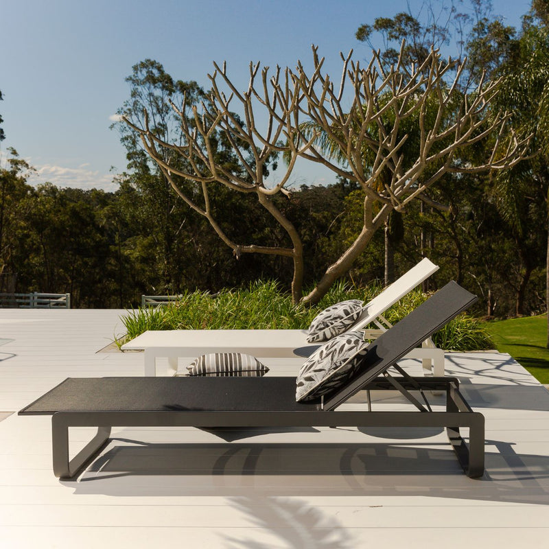 Two outdoor sun loungers, one black and one white, made from aluminium and textilene, are displayed from the side. The loungers are set in a sunny outdoor space with decorative pillows and a backdrop of greenery.