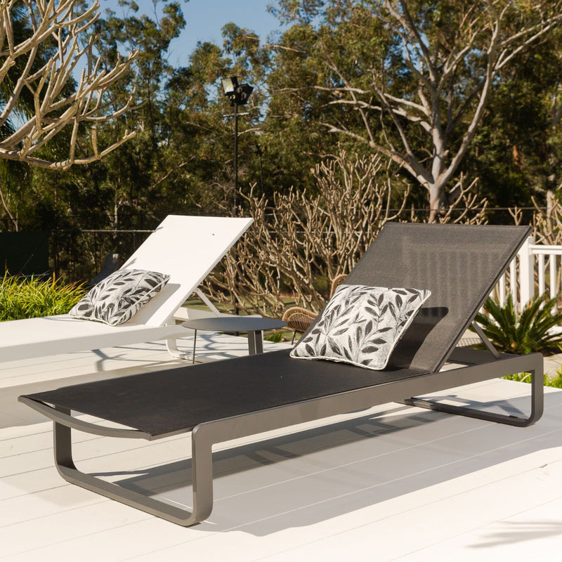 Two outdoor sun loungers, one black and one white, made from aluminium and textilene, are positioned in a sunny outdoor area. Decorative pillows adorn the loungers, and greenery is visible in the background.