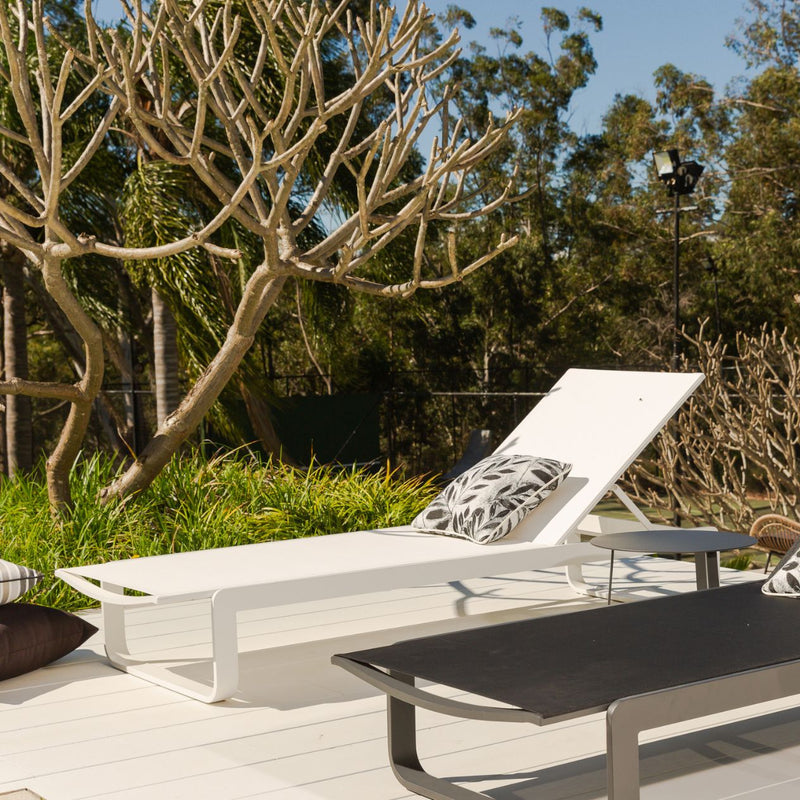 Two outdoor sun loungers, one black and one white, made from aluminium and textilene, are positioned in a sunny outdoor area. Decorative pillows adorn the loungers, and greenery is visible in the background.