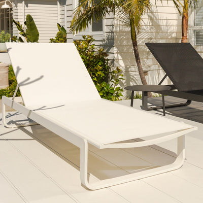 A white aluminium and textilene 'Verona' sun lounger positioned in a sunny outdoor area of someone's house, with the sun beaming down on it. A black sun lounger is partially cropped out of the frame, drawing focus to the white lounger.