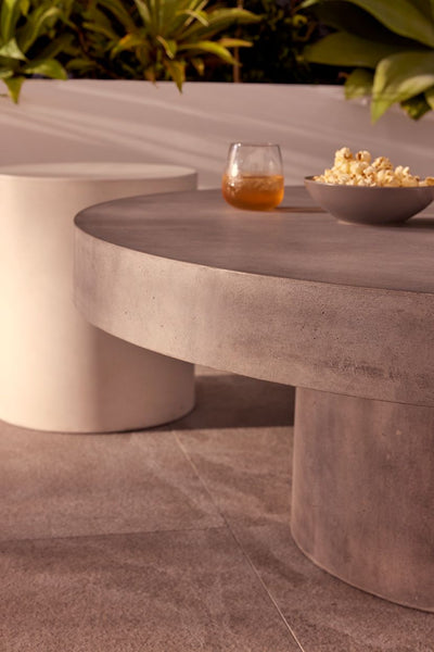 Zen round outdoor concrete coffee table, 90 cm, with a dark-grey weathered cement colour, in an outdoor area on stone tiling with some greenery in the background. On the table is popcorn and a glass of juice.
