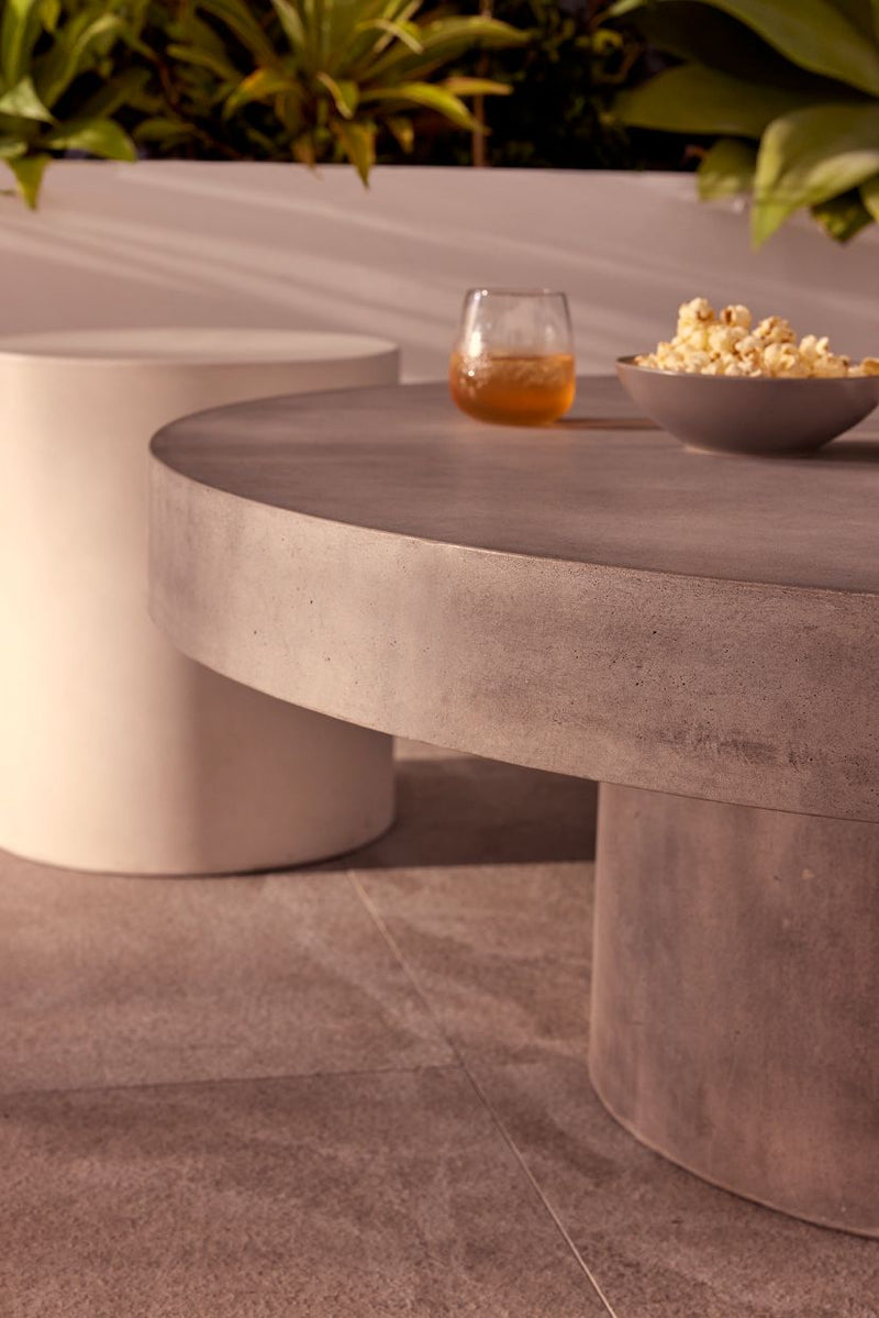 Zen round outdoor concrete coffee table, 90 cm, with a dark-grey weathered cement colour, in an outdoor area on stone tiling with some greenery in the background. On the table is popcorn and a glass of juice.