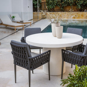 White Zen outdoor concrete round dining table with 4 dark grey rope outdoor chairs, set poolside with two pot plants on the table and beige sun loungers in the background, creating a stylish alfresco furniture setup.