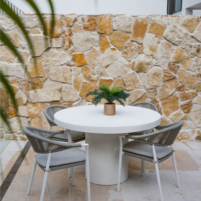 White Zen outdoor concrete round dining table with 4 light grey rope outdoor chairs, set against a sandstone wall with a pot plant on the table, offering a contemporary alfresco furniture look.