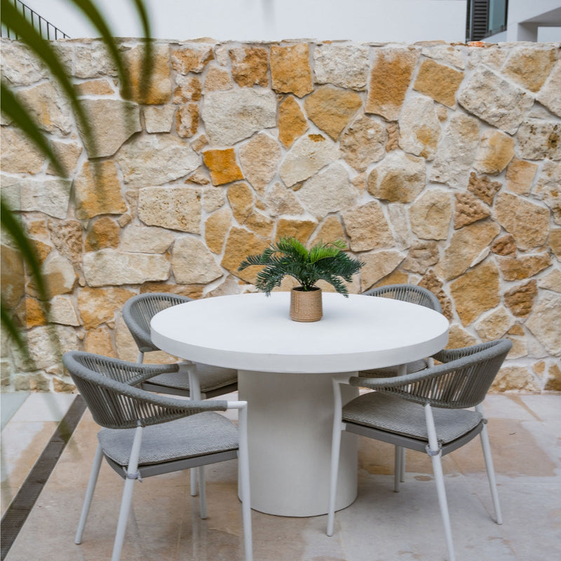 White Zen outdoor concrete round dining table with 4 light grey rope outdoor chairs, set against a sandstone wall with a pot plant on the table, offering a contemporary alfresco furniture look.