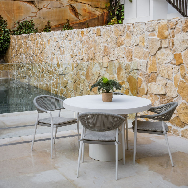 White Zen outdoor concrete round dining table with 4 light grey rope outdoor chairs, set against a sandstone wall with a pot plant on the table, and a pool visible in the background, creating a serene alfresco furniture setting.