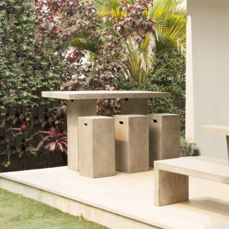 Outdoor setting featuring a sleek, weathered cement bar table with a minimalist design. Beside the table are 3 concrete bar stools. The table is 160 cm in length and is placed on a patio with greenery in the background, creating a serene atmosphere.