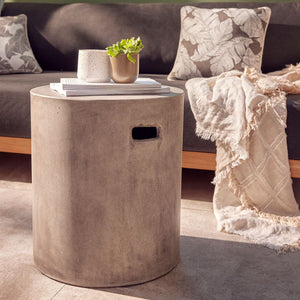 A cement-grey round outdoor stool crafted with glass-fibre-reinforced concrete, set in a sunny patio area. On the stool are two white books and two small pot plants. In the background, there is a timber outdoor lounge adorned with decorative throw pillows and a blanket.