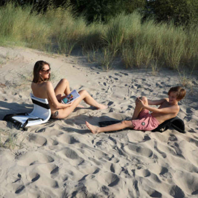 A mother and son relaxing on the beach using outdoor backrests. The mother is on a white 'Beach Buddy' backrest (which functions as a portable sun lounger), while the son uses a black one. Both are seated on the sand, enjoying the sunny weather and laughing together.