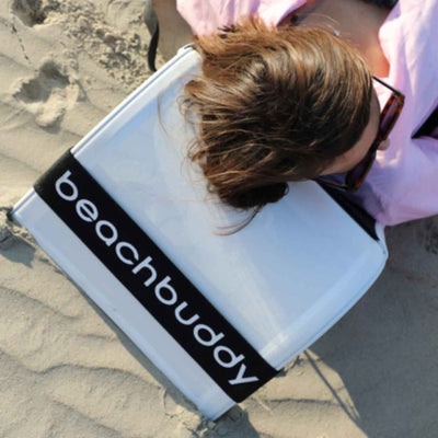 Close-up of a white 'Beach Buddy' backrest being used by a woman on the beach. The backrest is set on the sand, highlighting its design and comfort features for relaxation.