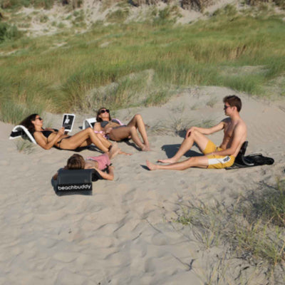 Four people in a circle on the beach: two girls and two guys. The girls are using white 'Beach Buddy' backrests (which function as portable sun loungers), and the guys are on black ones. They are all in swimwear, relaxing on the sand with some grass visible among the sand.