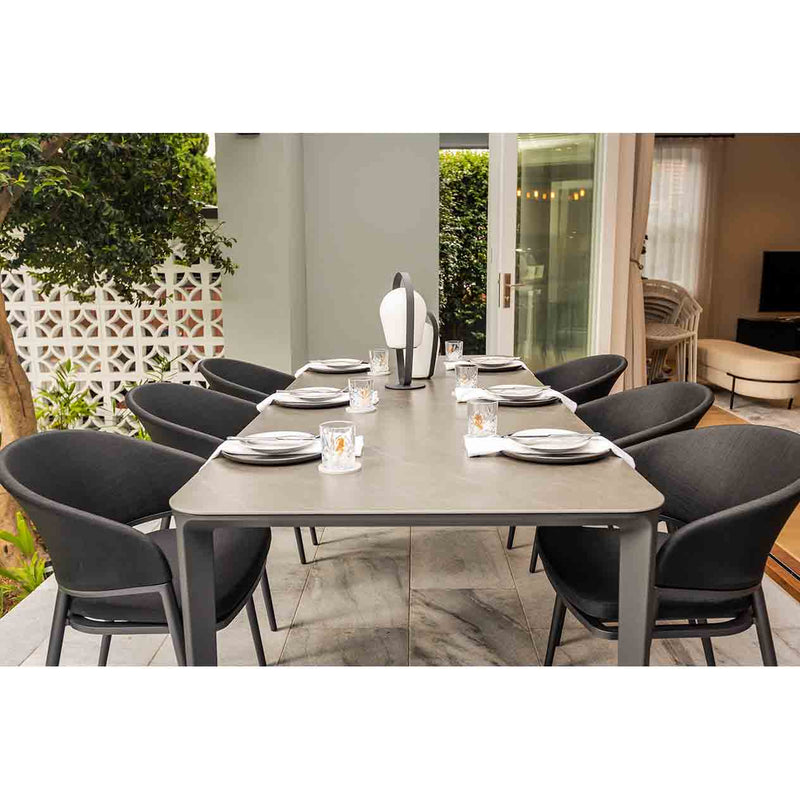 Six black aluminum Medea outdoor dining chairs with sleek zip-off cushion covers surrounding a rectangular outdoor dining table, making a set. The table is set with cutlery and outdoor lamps, positioned on a patio with a view of the backyard greenery and a glimpse of the indoor living area.