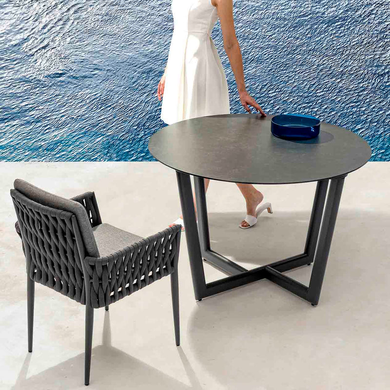 Black Colwood outdoor aluminium round dining table with ceramic glass top 110 cm accompanied by one matching outdoor chair, set poolside during summer. The alfresco furniture offers a stylish and comfortable spot for outdoor dining, perfect for enjoying warm weather by the pool.