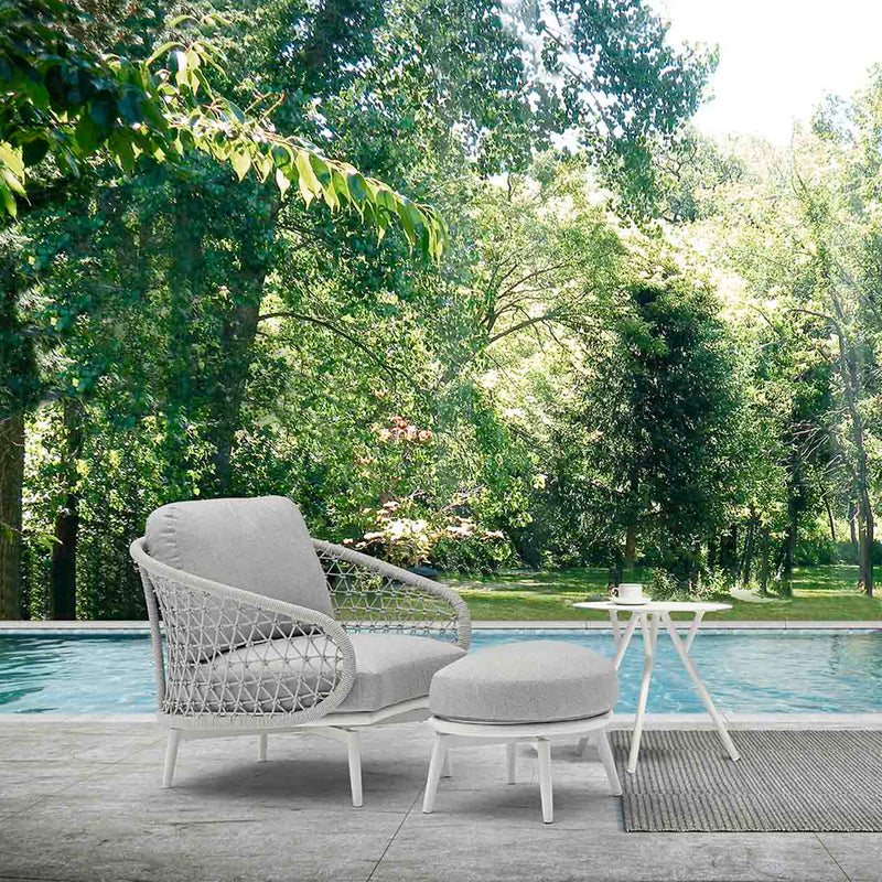 A tranquil outdoor living arrangement with a rope armchair, ottoman, and round aluminium coffee table, placed beside a pool surrounded by vibrant greenery and a scenic forest backdrop.