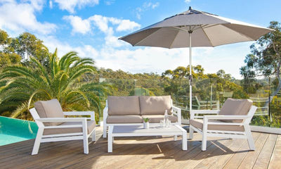 Light brown outdoor umbrella covering outdoor furniture on a wooden deck poolside, with greenery in the distance and blue skies, perfect for outdoor living.