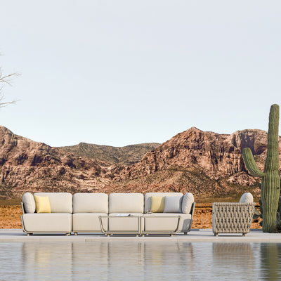 Outdoor modular sofa set in taupe with light sand-coloured cushions, complemented by yellow and grey accent pillows, paired with a matching rope-detailed armchair and a sleek coffee table in front, all set on a serene patio overlooking a desert landscape with rugged mountains and cacti.