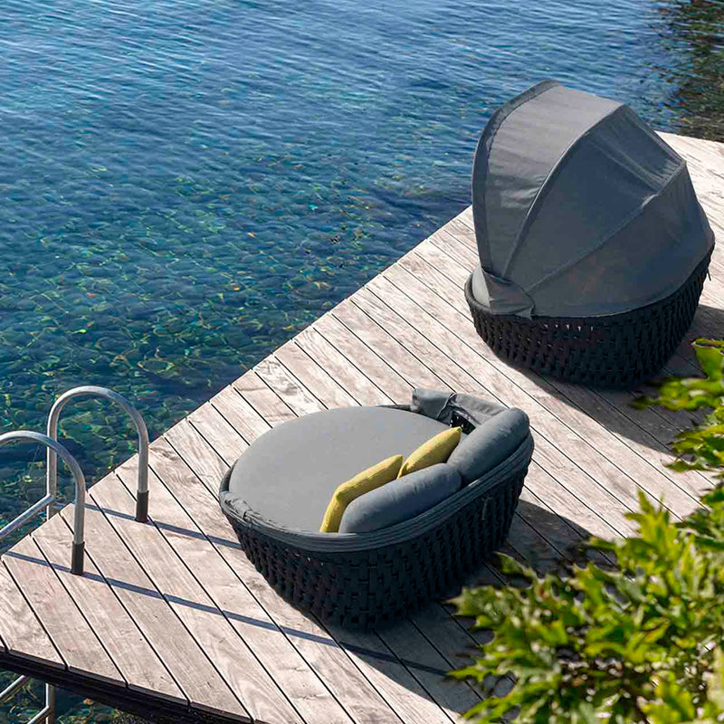 Two rope daybeds, one adorned with yellow pillows, set on a wooden deck overlooking serene water, with greenery peeking into the frame, perfect for outdoor living in summer.
