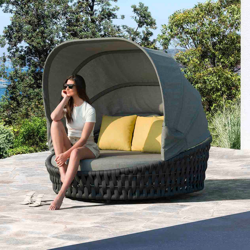 Rope daybed with two yellow pillows, a woman relaxing on it under the summer sun, surrounded by lush greenery, embodying the essence of outdoor living.