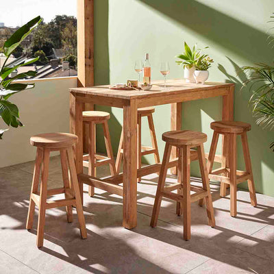The 200 cm 'Miles' outdoor bar table, a favourite among Remarkable Outdoor Living's wooden outdoor tables, is shown with five wooden stools arranged around it. This setup is positioned against a green wall in an outdoor area. The scene captures the table with wine and cheese, bathed in sunlight, with lush greenery surrounding the area.