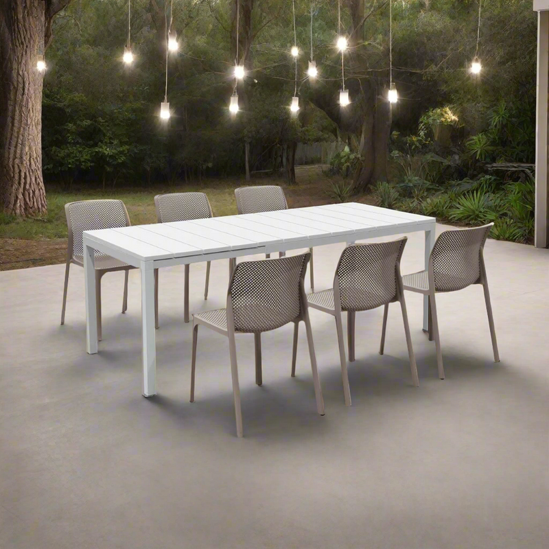Amidst a tranquil Australian bushland setting, illuminated by the gentle glow of hanging fairy lights, sits a contemporary dining set featuring a crisp white table paired with chic taupe chairs. The earthy, neutral tones of the chairs complement the natural textures of eucalyptus trees and native plants surrounding the deck. Lanterns and candles cast soft shadows, creating an intimate, magical feel as twilight descends. Accented with native floral arrangements—banksia, waratah, and eucalyptus leaves—this se