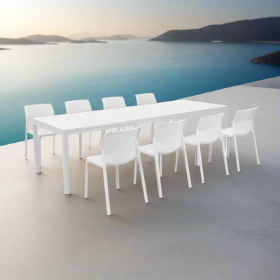Set against the backdrop of a sun-drenched Australian coastal terrace overlooking a glistening turquoise ocean, sits a sleek all-white dining set. The clean, crisp lines of the expansive white table and eight pristine chairs create an atmosphere of effortless sophistication. Around it, lush greenery in oversized pots and soft drapes gently billowing in the ocean breeze add a dreamy, whimsical touch. A scattering of soft beige cushions, textured ceramics, and a tray of fresh mangoes and sparkling drinks enha
