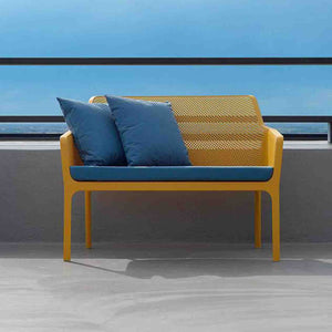 Rectangular bright-blue UV-stabilised 'Nardi Net' outdoor bench seat cushion on a mustard yellow resin Nardi outdoor bench seat, adorned with two blue pillows. The bench is displayed in a modern outdoor area with an ocean view in the background.