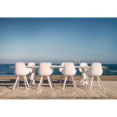 Front view of the Neverland alfresco furniture set with a Neolith® ceramic extension table set with four wine glasses and berries, paired with eight white resin dining chairs, positioned on a wooden path by the beach.