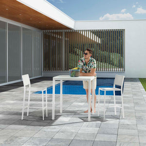 Two bar stools are positioned by a matching bar table, with a man leaning on the table, surrounded by fruit. This alfresco furniture setup is poolside at a modern house, captured on a sunny summer day, creating a perfect spot for relaxation and enjoyment.